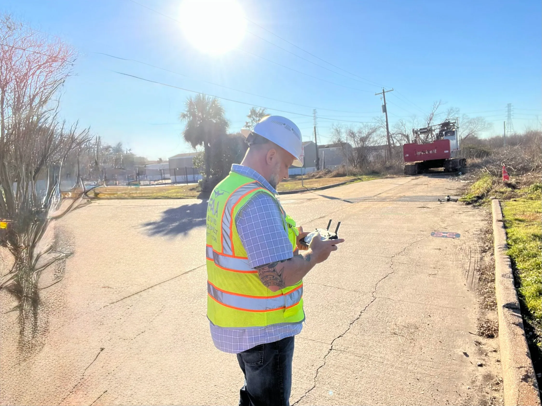Axis pilot in hard hat and high-visibility vest flying a drone over a cleared industrial site, tracked excavator working in the background.