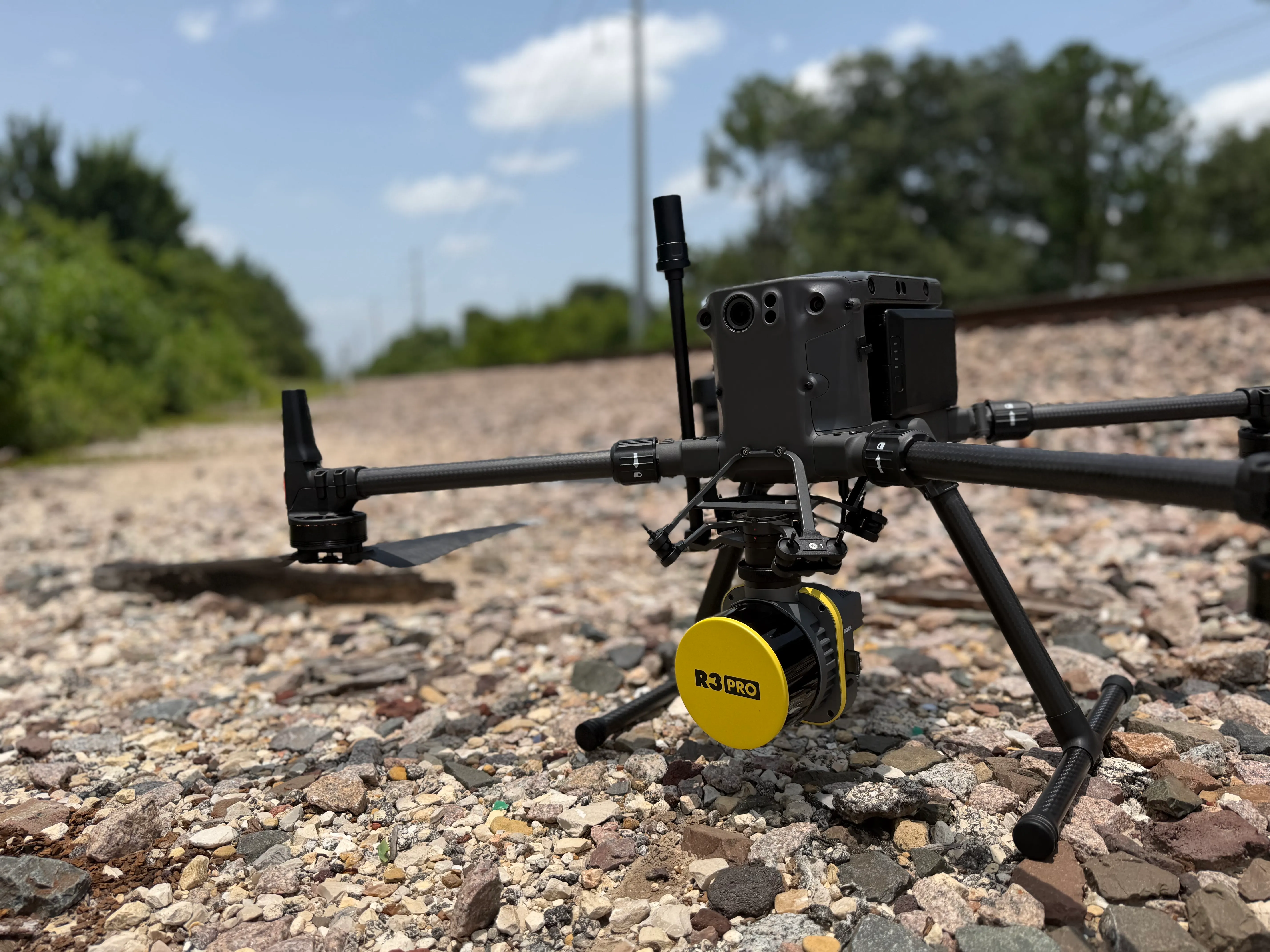 A UAS with a mounted R3 Pro LiDAR payload staged on gravel trackside ballast, framed against a soft-focus tree line and open sky.